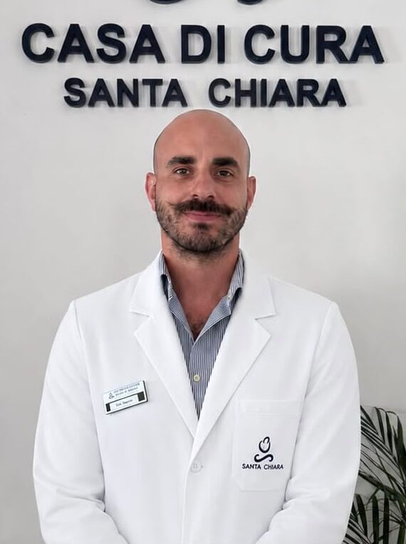 WhatsApp Image 2026-06-17 at 105610 - Casa di Cura Santa Chiara Male doctor in a white coat stands in front of a wall sign reading Casa di Cura Santa Chiara, in a clinic lobby.